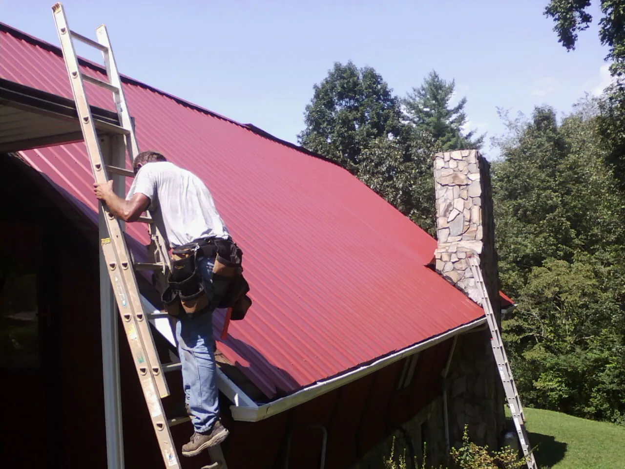 Roofer on a ladder beside a red metal roof and stone chimney — Soffit & Fascia Repair by Roofing Brothers in Taylorville
